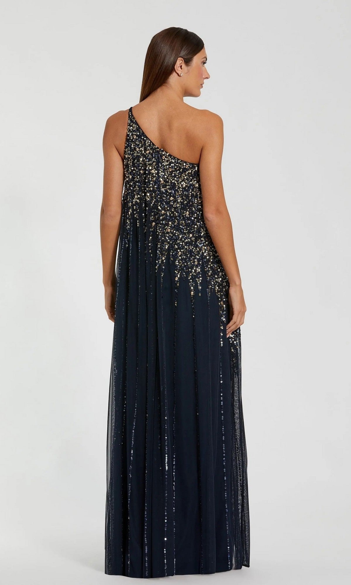 A woman stands facing away from the camera in the Mac Duggal 9224— a long, one-shoulder formal dress in dark blue, featuring gold and silver sequin beading, set against a plain background.